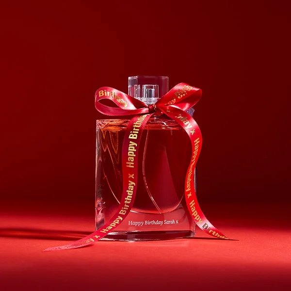 A perfume bottle tied with a red Happy Birthday ribbon and engraved with a personal message, on a red backdrop